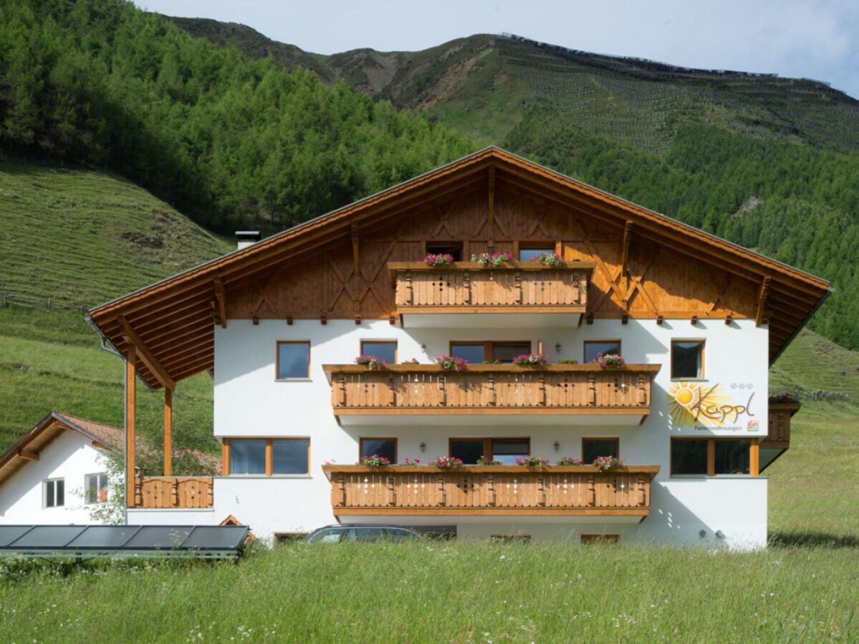 A multi-story building with balconies, a sloped roof, and a mountainous backdrop.