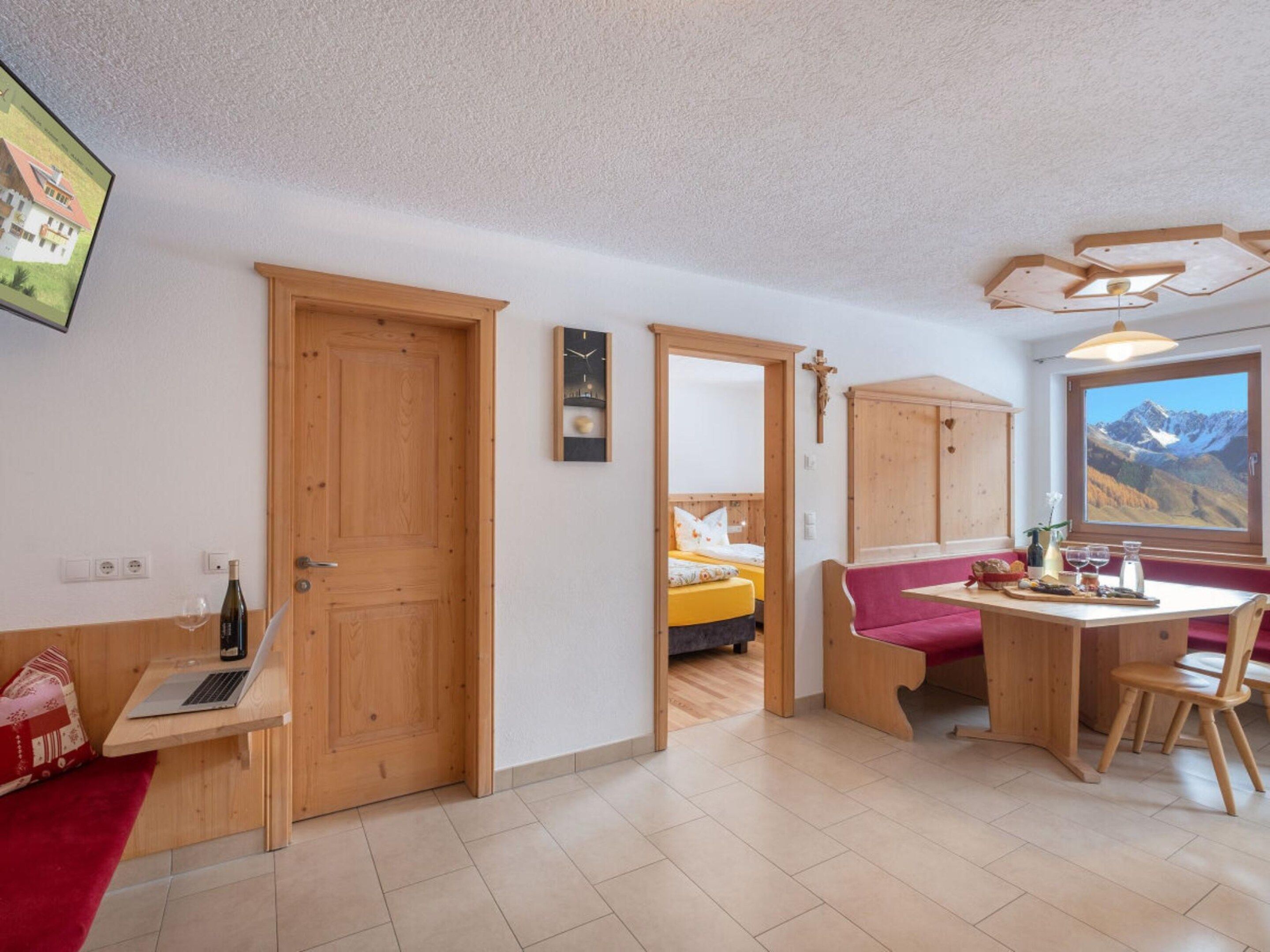 A room with a wooden door, a bed with a red headboard, a wooden table with chairs, a window with a mountain view, and a wall-mounted clock.