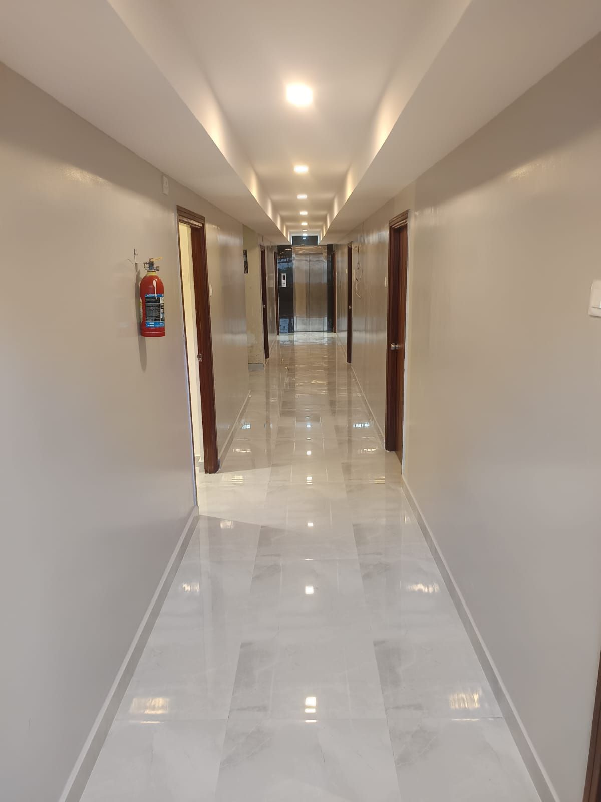 Un pasillo de hotel con un extintor de incendios montado en la pared, puertas a ambos lados y un piso de baldosas brillantes.