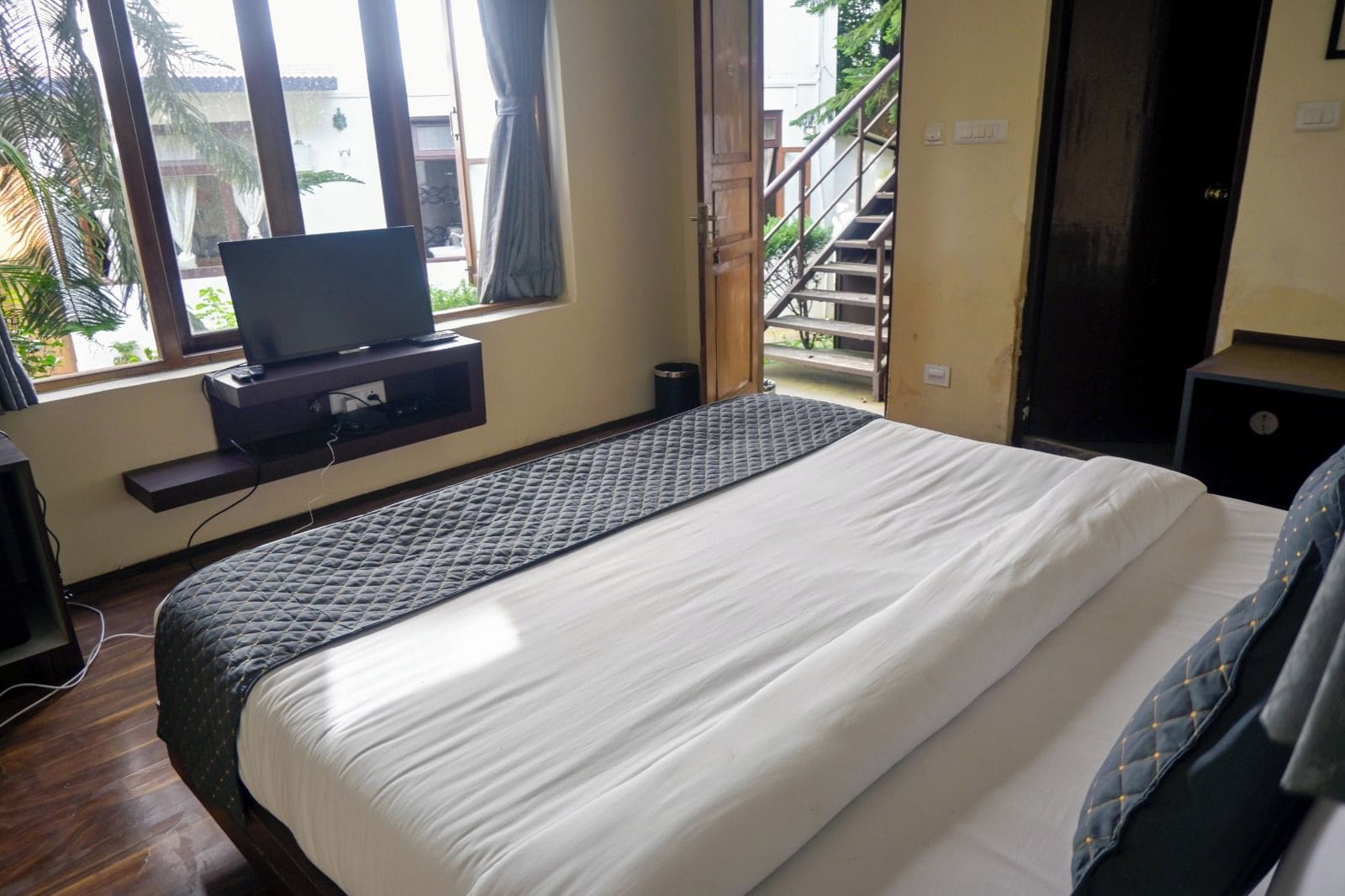 Un dormitorio con una cama, un televisor sobre un mueble bajo, una ventana con cortinas y vistas a una escalera exterior.