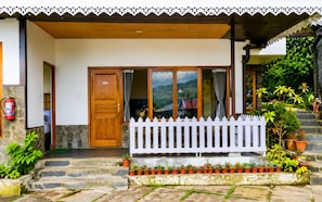 A wooden door with a number plate, a white picket fence, and potted plants in front of a building with a stone wall and a view of a mountain.