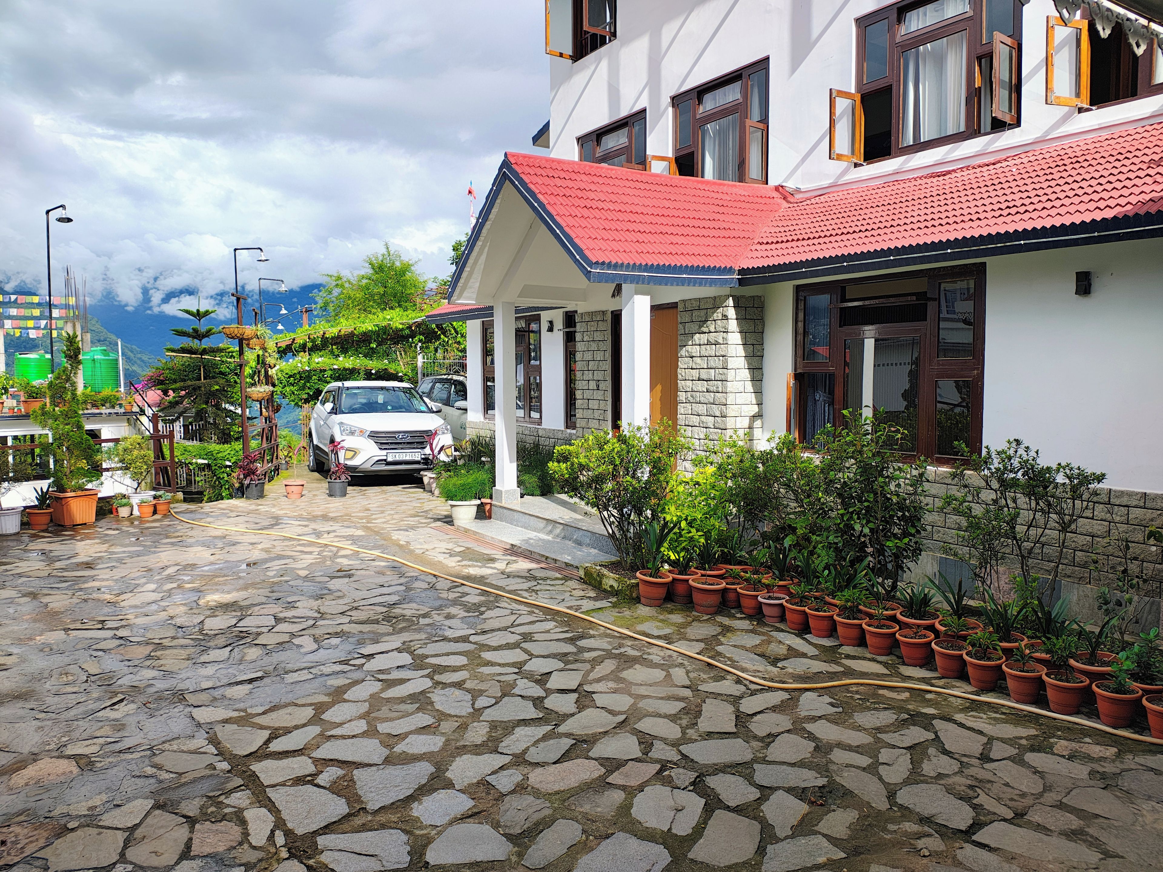 A paved driveway leading to a house with a red roof, surrounded by potted plants and a parked car.