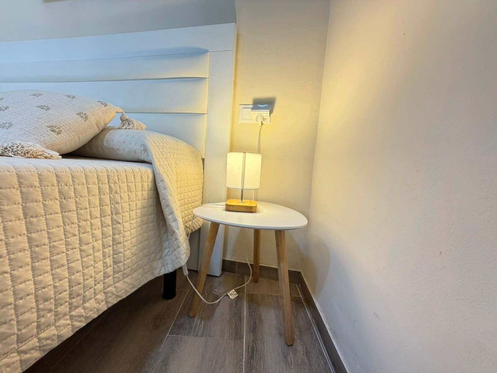 Una cama bien hecha con una colcha acolchada y almohadas, una mesita de noche con una lámpara, y un aplique de pared.