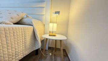 Una cama bien hecha con una colcha acolchada y almohadas, una mesita de noche con una lámpara, y un aplique de pared.
