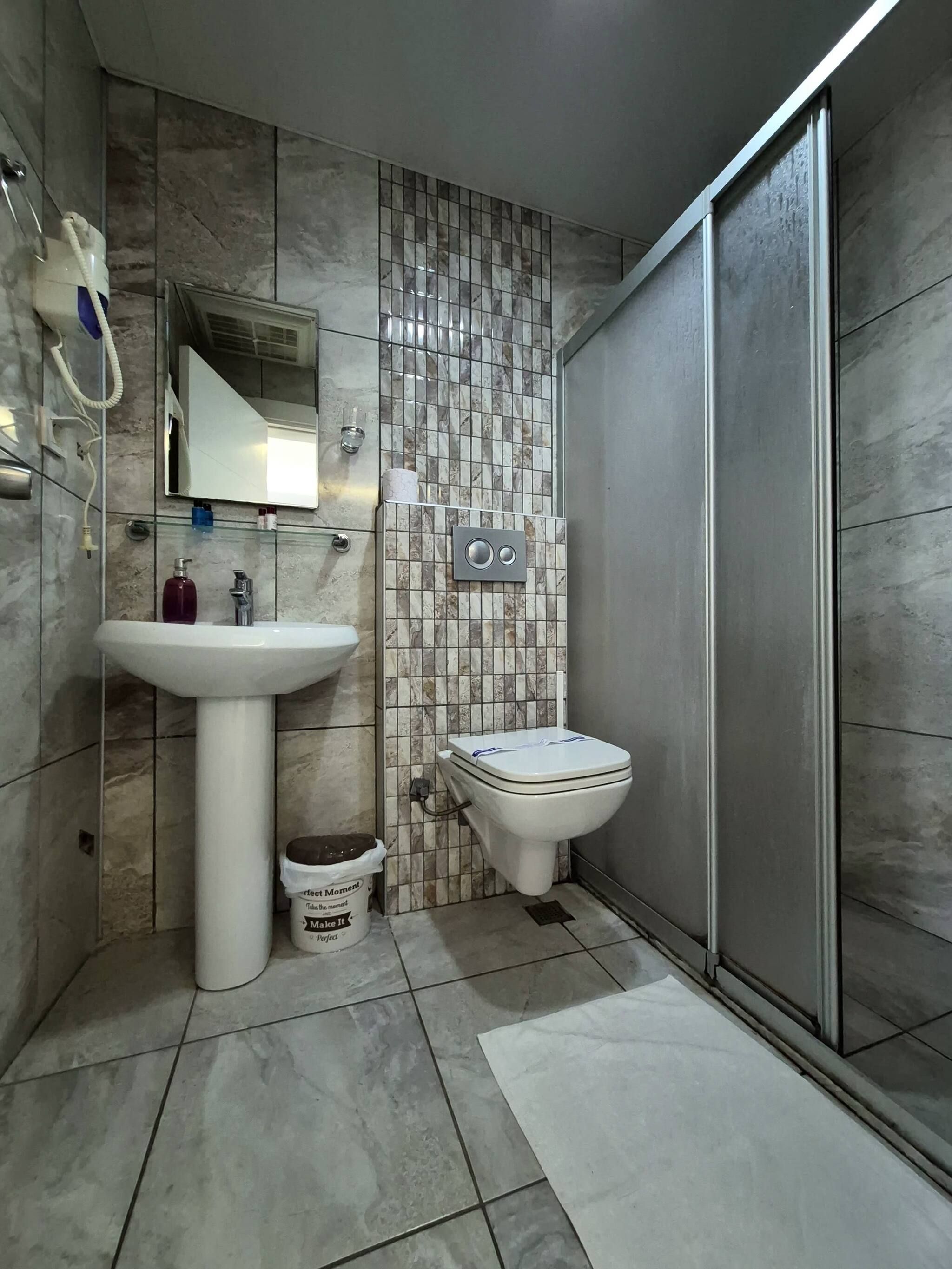 A bathroom with a pedestal sink, a toilet, a shower area, and a tiled wall.
