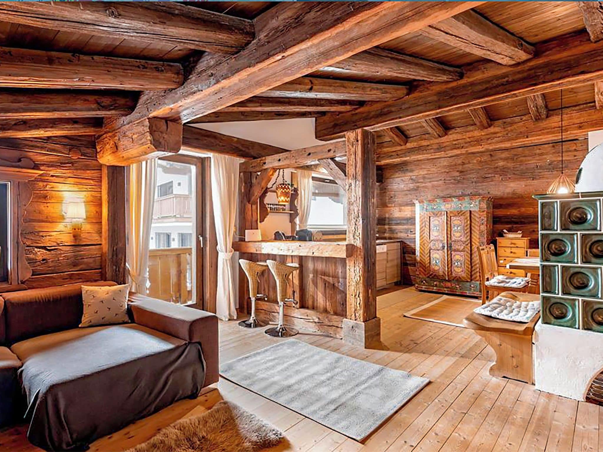 A rustic cabin interior with exposed wooden beams, a sofa, a dining area, and a kitchen with a decorative stove.