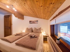 A bedroom with a wooden ceiling, a bed with checkered bedding, a nightstand with a lamp, and a window with a view. - Manharthof by Interhome (Kirchberg in Tirol)
