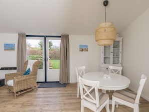 A room with a dining table, white chairs, a wicker chair, a cabinet, and a view through a glass door. - Holiday Home in Renesse With Swimming Pool (Renesse)