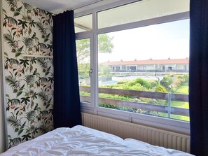 A bedroom with a large window, a bed, and a view of a building and greenery outside. - Apartment in Westkapelle With Wi-fi (Westkapelle)