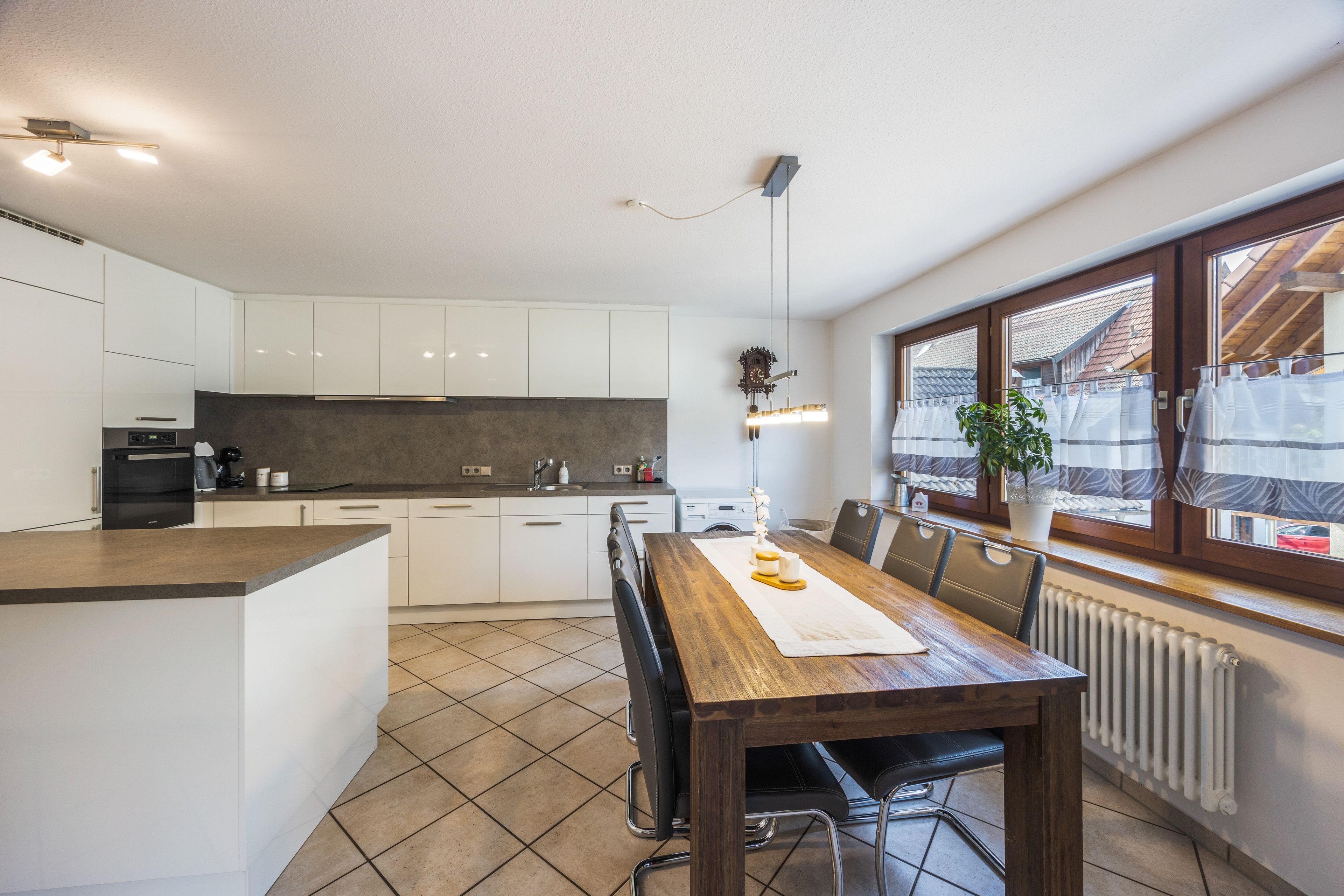 Eine moderne Küche mit weißen Schränken, einem Holztisch und Blick auf eine Wohngegend durch ein großes Fenster.