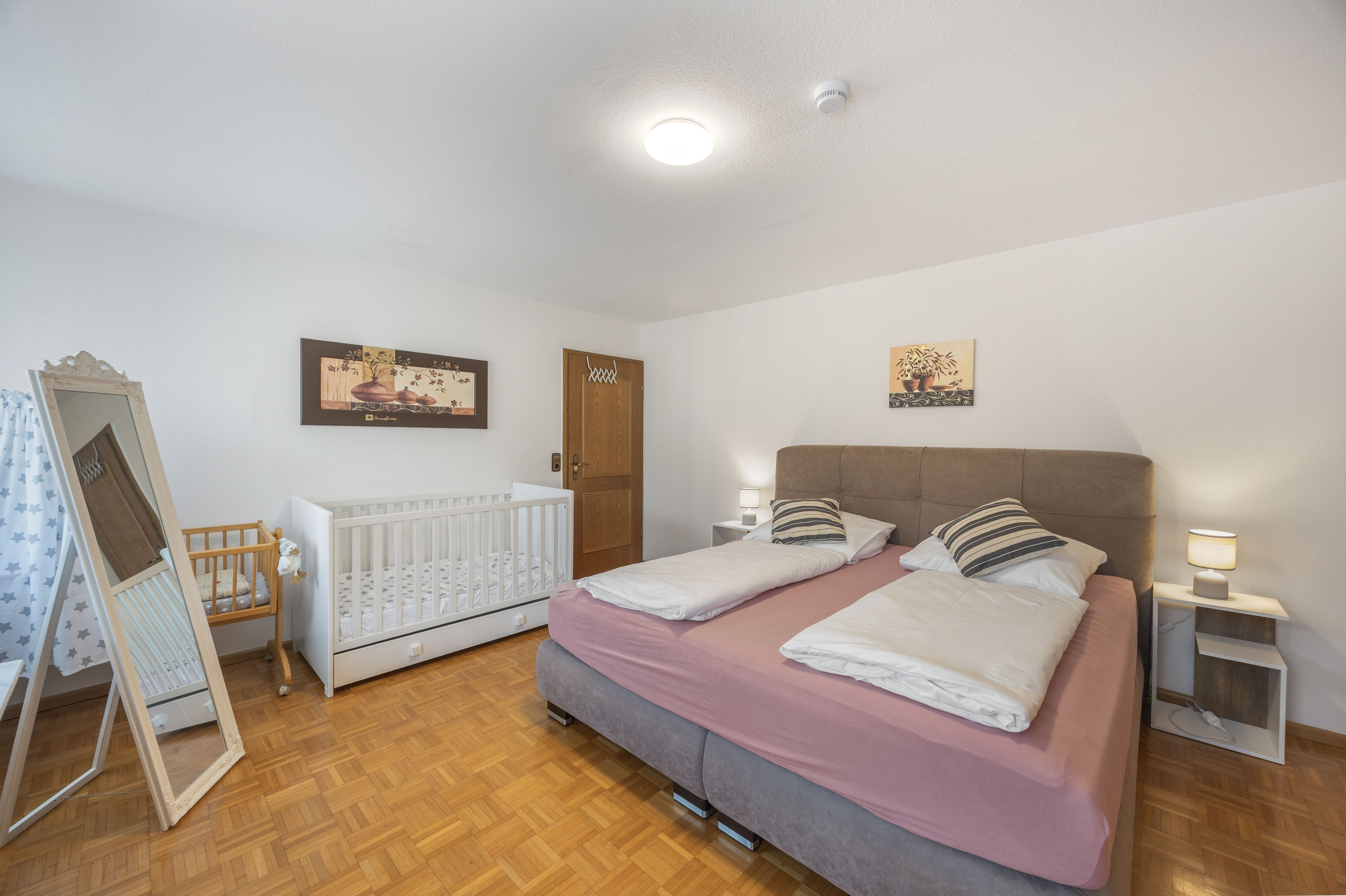Ein Schlafzimmer mit einem Doppelbett, einem Babybett, Holzboden und einer Tür.
