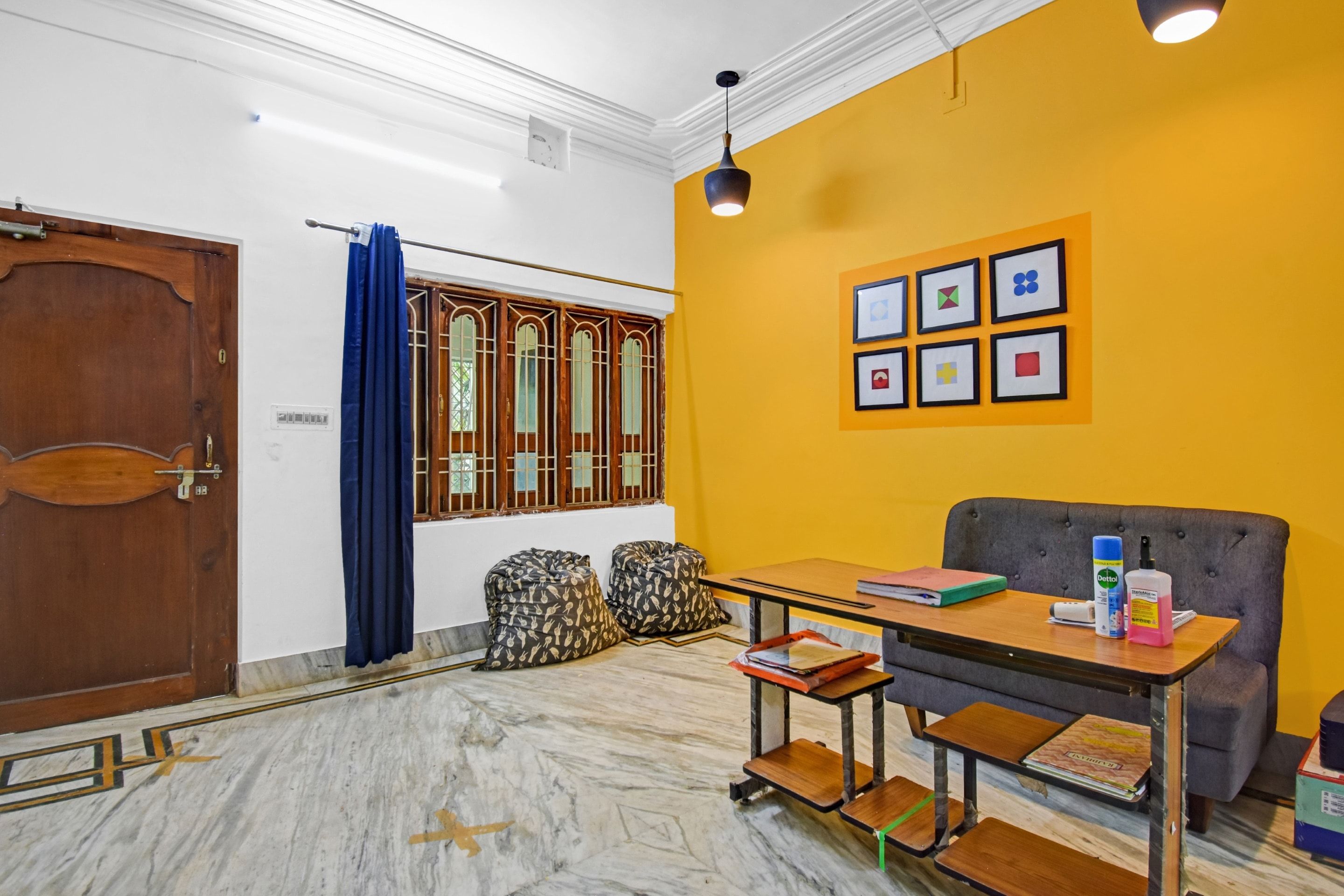 Una habitación con puerta de madera, cortina azul, ventana con barrotes de madera, pared amarilla con cuadros, sofá gris, mesa de madera con libros y una botella, y suelo de mármol.
