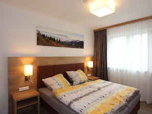 A bedroom with a bed, two bedside lamps, a nightstand, a painting on the wall, and a window with curtains. - H&P Residence Grafenmatt Type B (Feldberg)
