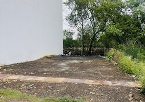 Una zona pavimentada con un muro blanco a un lado y una arbolada con césped al otro.