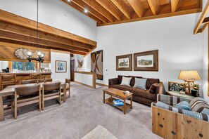 A spacious living area with a wooden ceiling, a dining table with chairs, a sofa, a coffee table, and a lamp. - Pine Views • Shared Pool + Spa | Steps to Northstar Shuttle | Northstar Pines (Truckee)