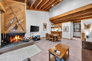 A cozy living room with a fireplace, wooden beams, a TV, and a dining area. - Pine Views • Shared Pool + Spa | Steps to Northstar Shuttle | Northstar Pines (Truckee)