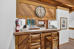 A kitchen with a wooden bar, a large wall clock, and a fire extinguisher. - Pine Views • Shared Pool + Spa | Steps to Northstar Shuttle | Northstar Pines (Truckee)