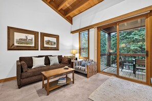 A cozy living room with a brown sofa, a wooden coffee table, and a bookshelf. There are two framed pictures on the wall, and a sliding glass door leads to an outdoor deck with a view of trees. - Pine Views • Shared Pool + Spa | Steps to Northstar Shuttle | Northstar Pines (Truckee)