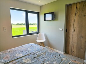 A bedroom with a large window, a bed with a floral bedspread, a white chair, a wooden door, and a mounted television. - Holiday Home With 1 Bedroom (Aagtekerke)