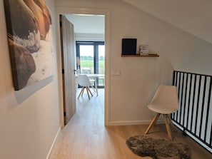 A hallway with a wooden floor, a white chair, a black and white striped headboard, and a view of a balcony with a table and chairs. - Holiday Home With 1 Bedroom (Aagtekerke)