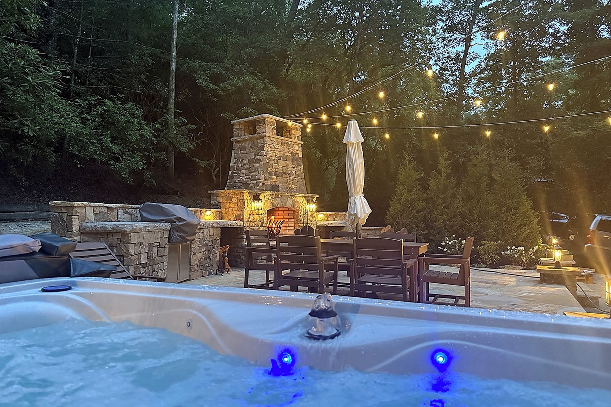 A hot tub with a stone fireplace and outdoor seating, surrounded by trees and string lights.