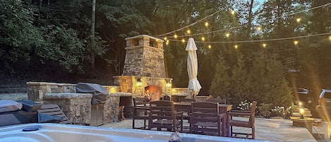 A hot tub with a stone fireplace and outdoor seating, surrounded by trees and string lights.