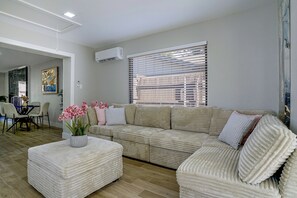 A living room with a beige sofa, a coffee table with a vase of pink flowers, and a window with blinds. - Cozy Home Base Btwn Dallas & Fort Worth (Bedford)