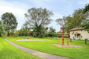 Ein Spielplatz mit Schaukeln und einer Kletterlandschaft, umgeben von Gras und Bäumen.