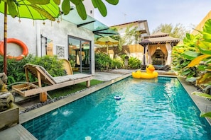 Une piscine dotée d’une terrasse en bois et de chaises longues, entourée d’une végétation luxuriante et d’une maison avec une porte en verre.