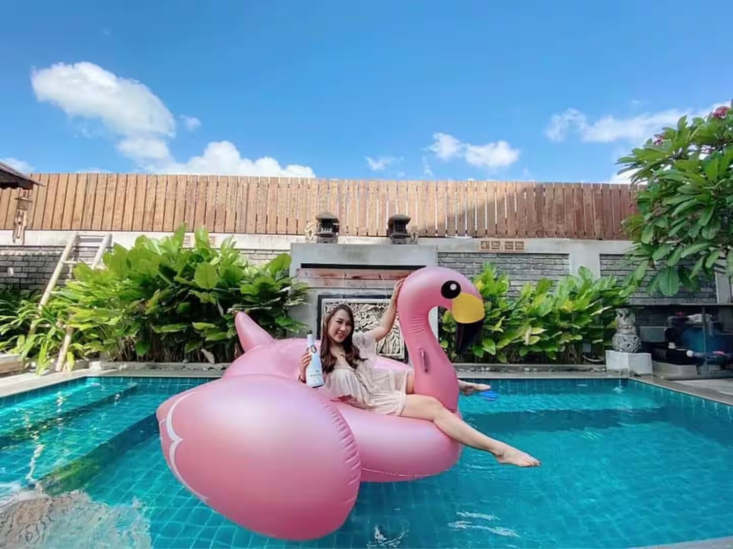 A person sitting on a pink inflatable flamingo in a swimming pool with a wooden fence and greenery in the background.