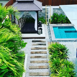 A pool area with a wooden deck leading to a covered seating area, surrounded by lush greenery.