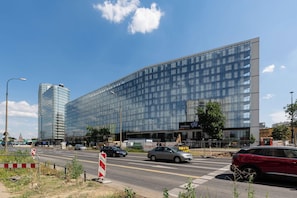 Ein modernes Gebäude mit Glasfassade, eine Straße davor mit Autos und einem roten Fahrzeug, und ein klarer, blauer Himmel.