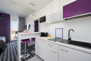 A compact hotel room with a kitchenette, a bed, a window with curtains, and a view of the cityscape. - Warsaw | Kasprzaka 31 | Modern Studio | Balcony | Air Conditioning (Warszawa)