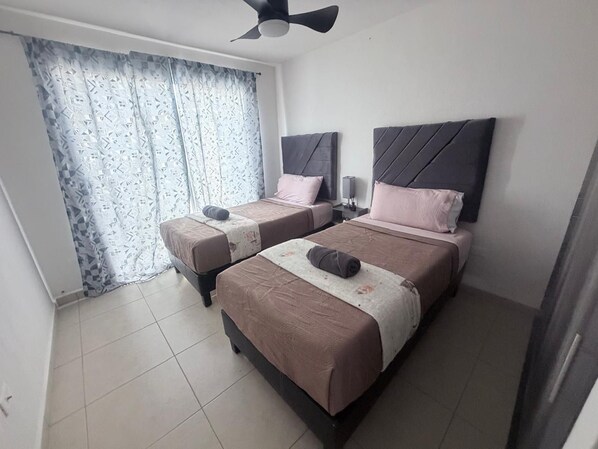 A hotel room with two beds, a ceiling fan, and a window with patterned curtains.