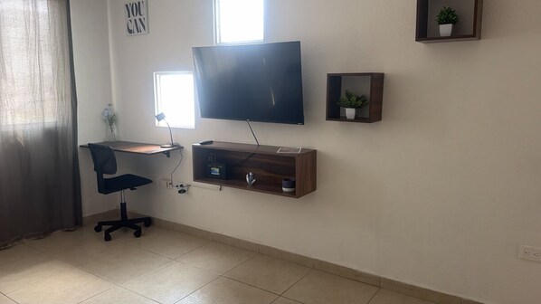 A room with a desk, chair, TV, and shelves.