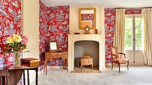 Una habitación con chimenea, un espejo, una silla y una mesa con un jarrón de flores.