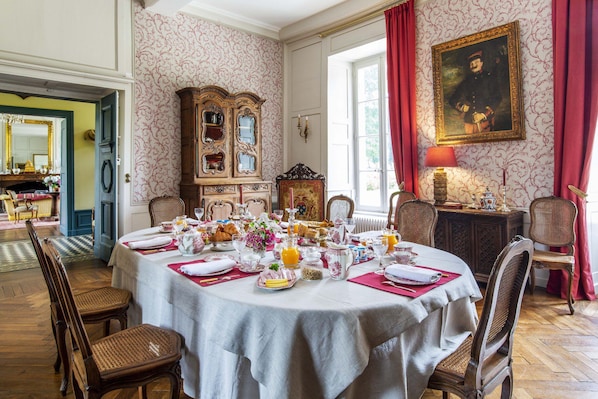 Comedor con una mesa redonda dispuesta para una comida, rodeado de muebles antiguos y un cuadro grande.