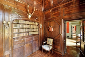 Una habitación con paneles de madera, una estantería llena de libros, un sillón con tapicería floral y una cabeza de ciervo montada en la pared.