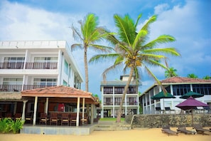 Un hotel frente a la playa con edificios de varios pisos, una zona de estar cubierta y una playa de arena con sillones de descanso.