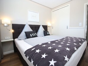Ein ordentlich bezogenes Bett mit einer Decke mit Sternenmuster, zwei Kissen mit Aufdruck, ein Kopfteil, Nachttischlampen und ein gerahmtes Bild an der Wand.