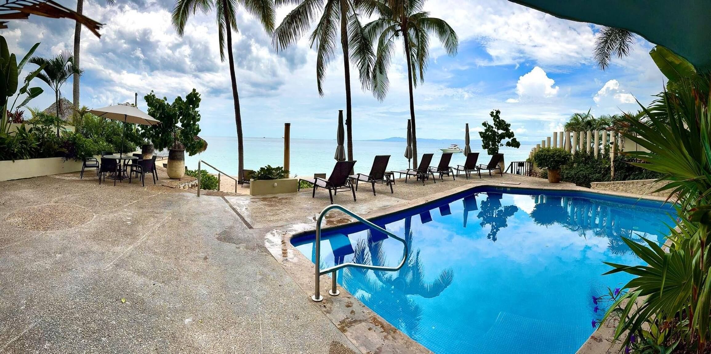 Una piscina con sillas de descanso y sombrillas, rodeada de palmeras y vista al mar.