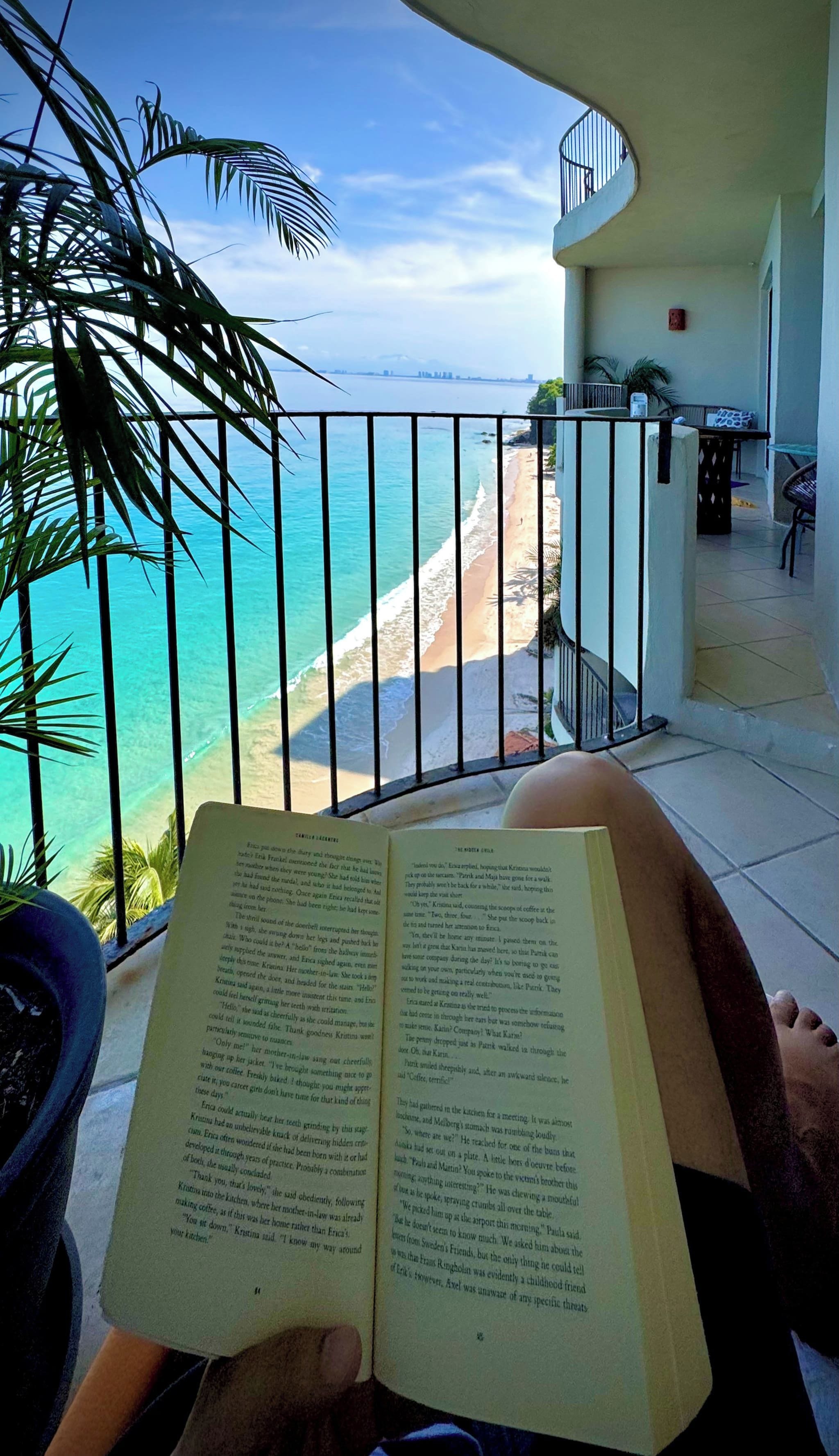 Una persona leyendo un libro con vista a una playa y al océano, a través de la barandilla de un balcón.