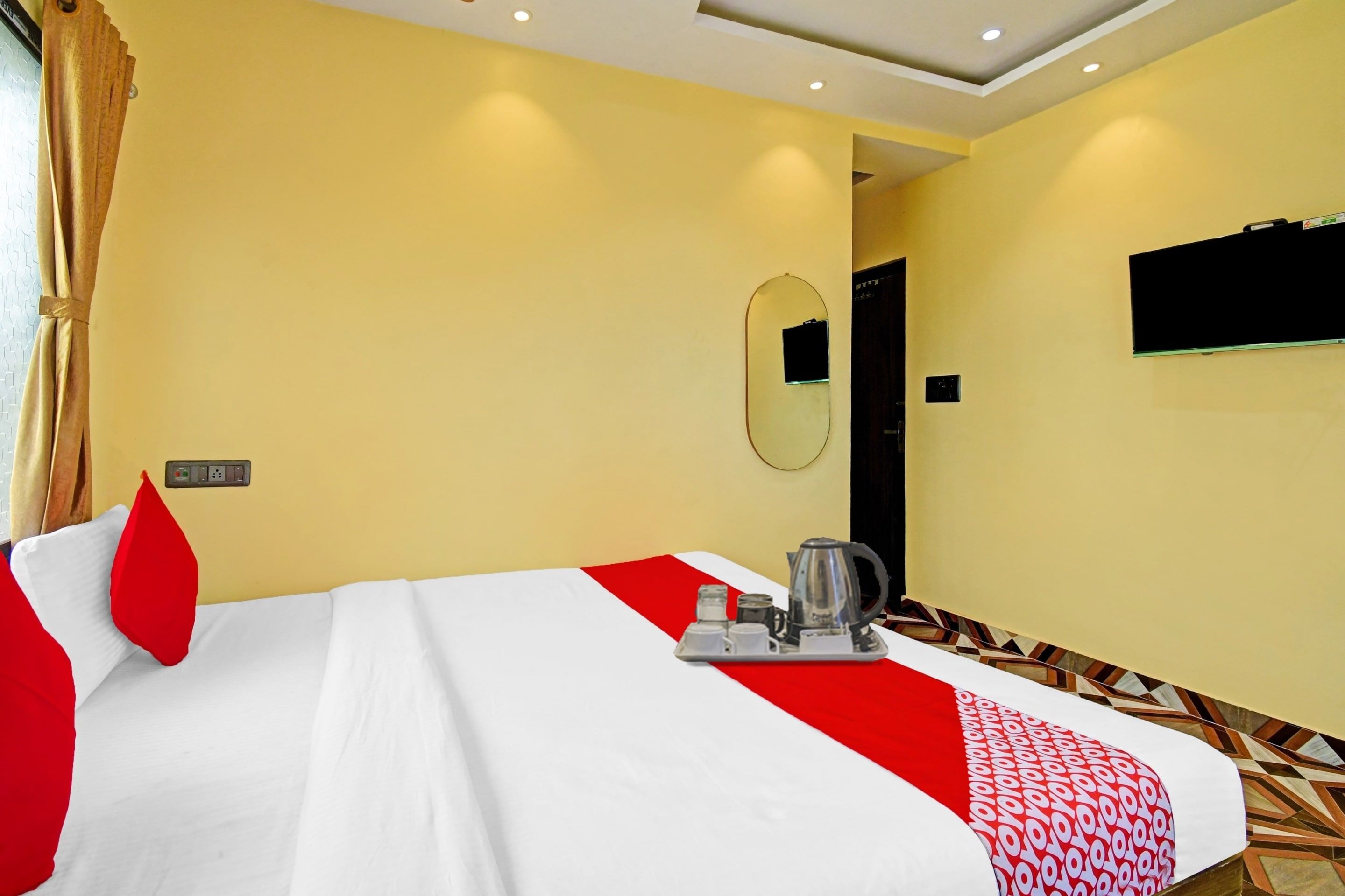 A hotel room with a bed, a bedside table with a kettle, a mirror, and a TV on the wall.