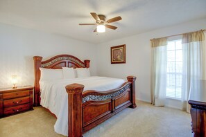 Um quarto com uma cama de madeira com dossel, uma mesa de cabeceira com abajur, um ventilador de teto, uma janela com cortinas e um quadro na parede.