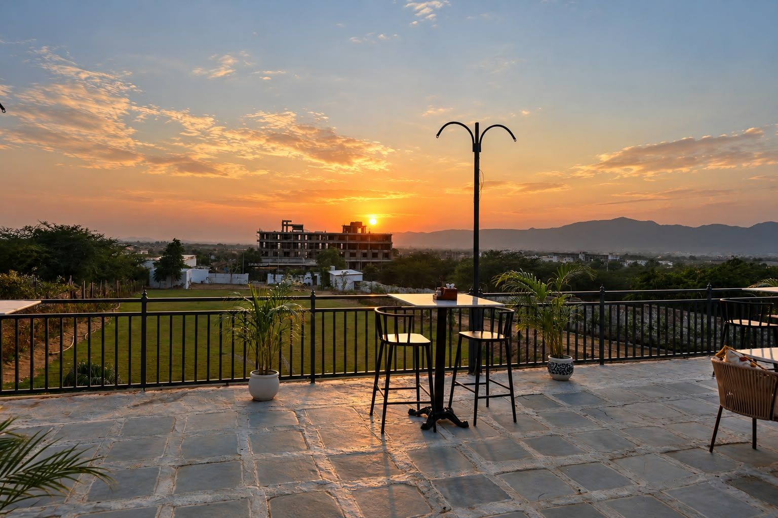 Una terraza con una mesa y sillas, una planta en maceta y vistas a un edificio y paisaje al atardecer.