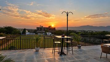 Una terraza con una mesa y sillas, una planta en maceta y vistas a un edificio y paisaje al atardecer.