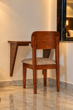 A wooden chair with a cushioned seat and backrest, positioned in front of a marble floor and a wall-mounted wooden shelf. - Rahvaas Hotel & Resorts (Pushkar)