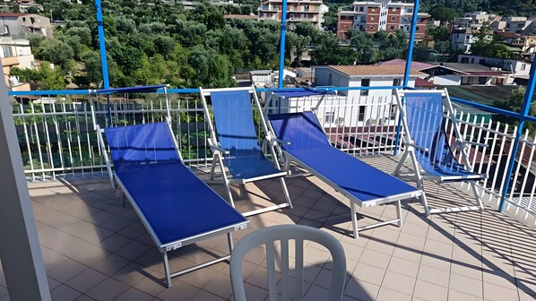 Ein Balkon mit blauen Liegestühlen und einem weißen Stuhl mit Blick auf eine Wohngegend mit Bäumen und Gebäuden.