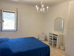 Ein Schlafzimmer mit Bett, Kommode, Spiegel und einem Fenster mit Blick ins Freie.