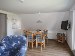 Ein Zimmer mit Esstisch, Stühlen, einem blauen Sessel, einem Fernseher und einem Heizkörper.